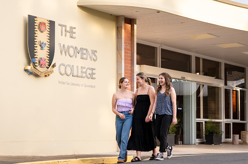 Students walk outside a building, with a sign in the background saying "The Women's College within The University of Queensland".